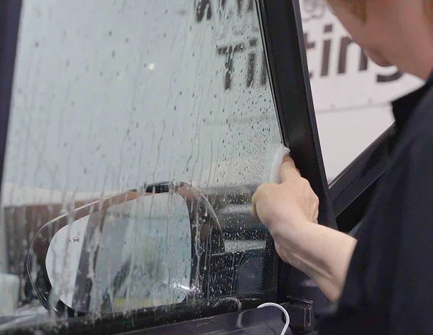 Close-up of a person applying window tint film to a wet car window, using a tool to press the film into the corner near the frame.