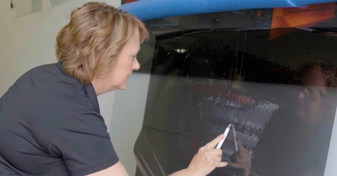 A woman uses a white applicator tool to smooth and press tinted film onto a car’s rear windshield, with a blue foam guard protecting the window edge.