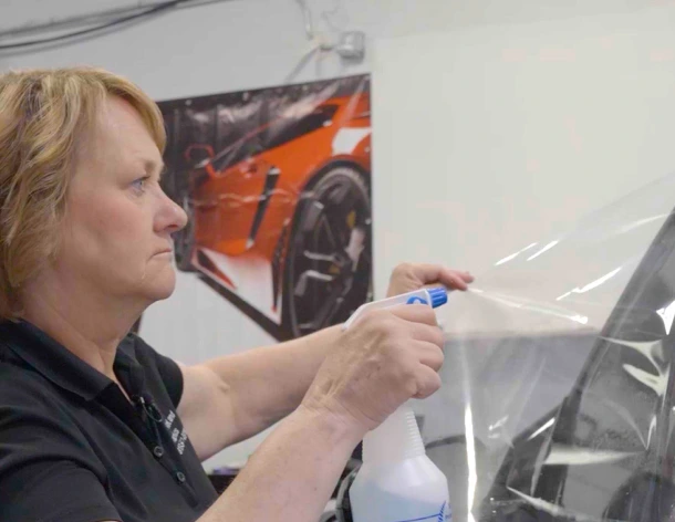 A woman applies a spray solution to a large clear sheet of tint film while installing it on a car window, with a vivid orange sports car poster in the background.
