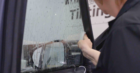 A person uses a tool to press down window tint film on a wet vehicle window, smoothing it along the frame edge inside a tinting shop.
