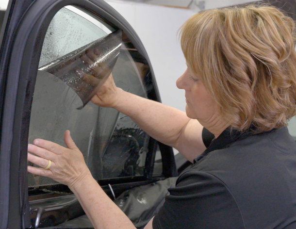 A woman carefully aligns and presses a sheet of tint film onto a wet car door window during the window tinting process in a workshop setting.