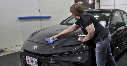 A woman uses a blue microfiber cloth to wipe down the hood of a dark gray Toyota sedan in a detailing garage. The car has a front license plate and detailing supplies are visible in the background.