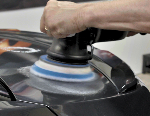 Close-up of a person using a dual-action polisher with a blue and white foam pad to buff the surface of a black vehicle, likely applying polish or preparing for a ceramic coating.