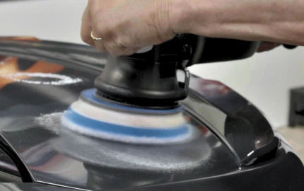 Close-up of a person using a dual-action polisher with a blue and white foam pad to buff the surface of a black car panel, creating a swirl of polishing compound.