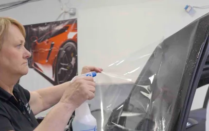 A woman is spraying liquid onto a large sheet of clear window tint film as she applies it to a vehicle window in a garage with a sports car poster in the background.