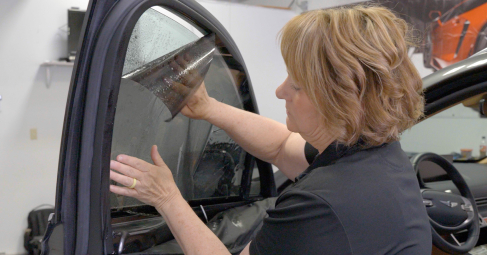 A woman is carefully installing window tint film on a car door’s side window, aligning the tinted sheet and smoothing it with her hands.