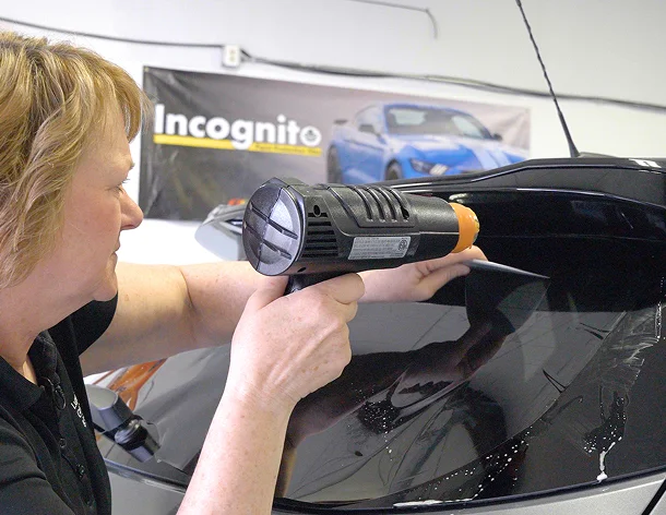 A woman uses a heat gun to apply or adjust window tint film on the rear windshield of a vehicle, with a banner reading “Incognito” and a blue car in the background.
