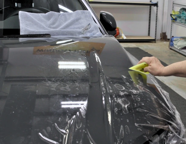 A person uses a yellow squeegee to apply a paint protection film (PPF) to the hood of a dark-colored car, with visible soap and water solution beneath the film. A towel covers part of the windshield for protection during the installation.