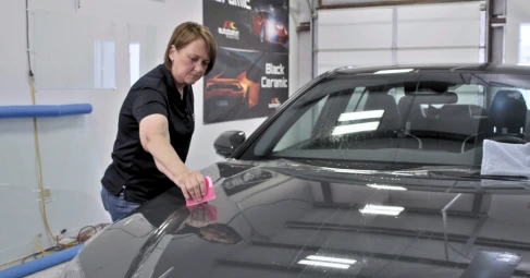 A woman uses a pink squeegee to smooth out a protective film on the hood of a dark-colored car inside a detailing garage. The car’s surface appears wet, and a towel is placed on the windshield; posters on the wall advertise ceramic coatings.