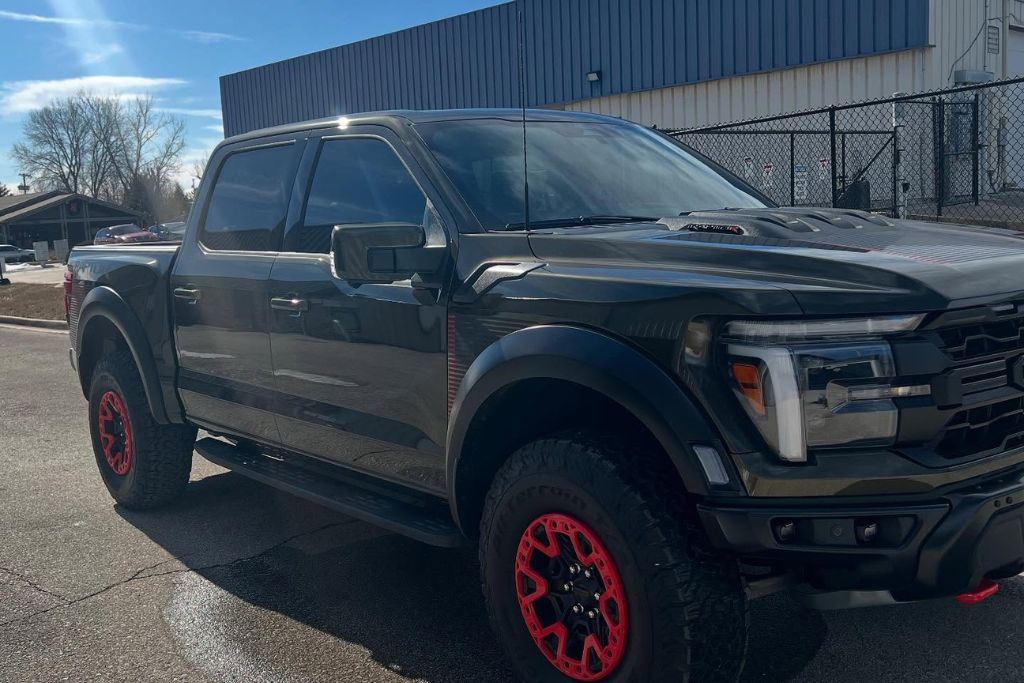 A black Ford Raptor pickup truck with red-accented wheels is parked in the sunlight, showing off its glossy, freshly ceramic-coated finish. The truck is outside an industrial building with a fence and blue-roofed structure in the background.