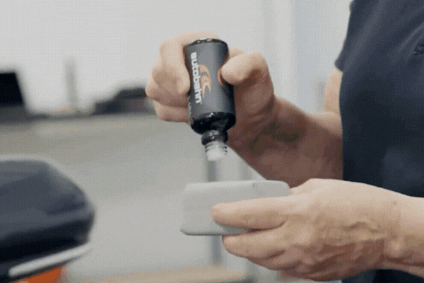 A person applies ceramic coating by pouring liquid from a black bottle labeled "W6Pro" onto a gray applicator pad, preparing to treat a vehicle's surface. The scene takes place in a workshop environment.
