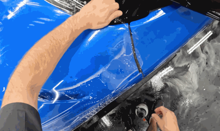 A technician aligns and stretches a clear protective wrap across a blue car's body panel. The surface is wet to help the film adhere without bubbles