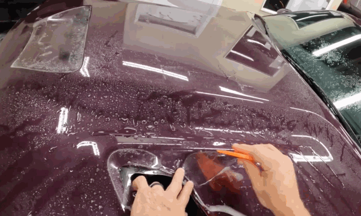Hands install a paint protection film on a maroon car’s hood, carefully trimming around a raised vent using an orange squeegee. Water droplets cover the surface during the process
