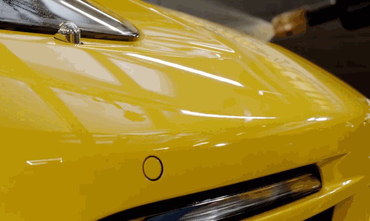A close-up of a yellow car’s front bumper shows the paint protection film being smoothed out, revealing a glossy, seamless finish under bright shop lighting