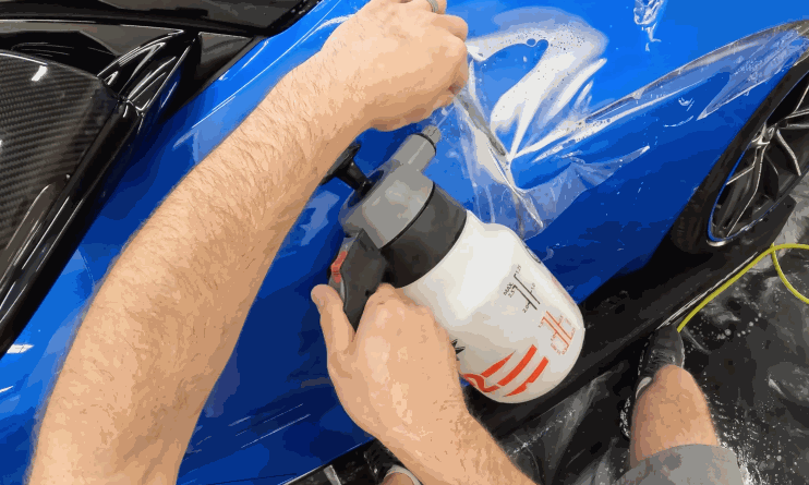 A person uses a spray bottle to apply a protective clear film to the fender of a glossy blue sports car. The film is being carefully positioned over the paint to ensure smooth adhesion