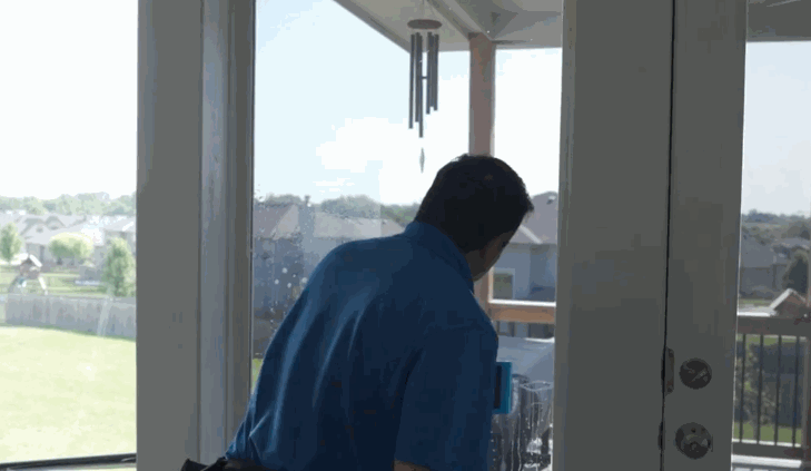 A window tint installer uses a squeegee to finish applying film on a tall glass door, with a ladder nearby and a view of a suburban neighborhood outside.