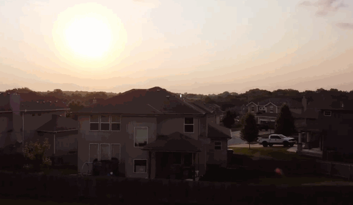 A time-lapse of the sun setting over a suburban neighborhood, showing sunlight gradually dimming across rooftops and trees and a home with window tint