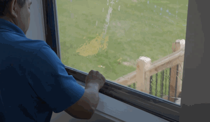 A technician in a blue shirt installs a clear protective window film on a residential window from inside the house, overlooking a grassy yard.