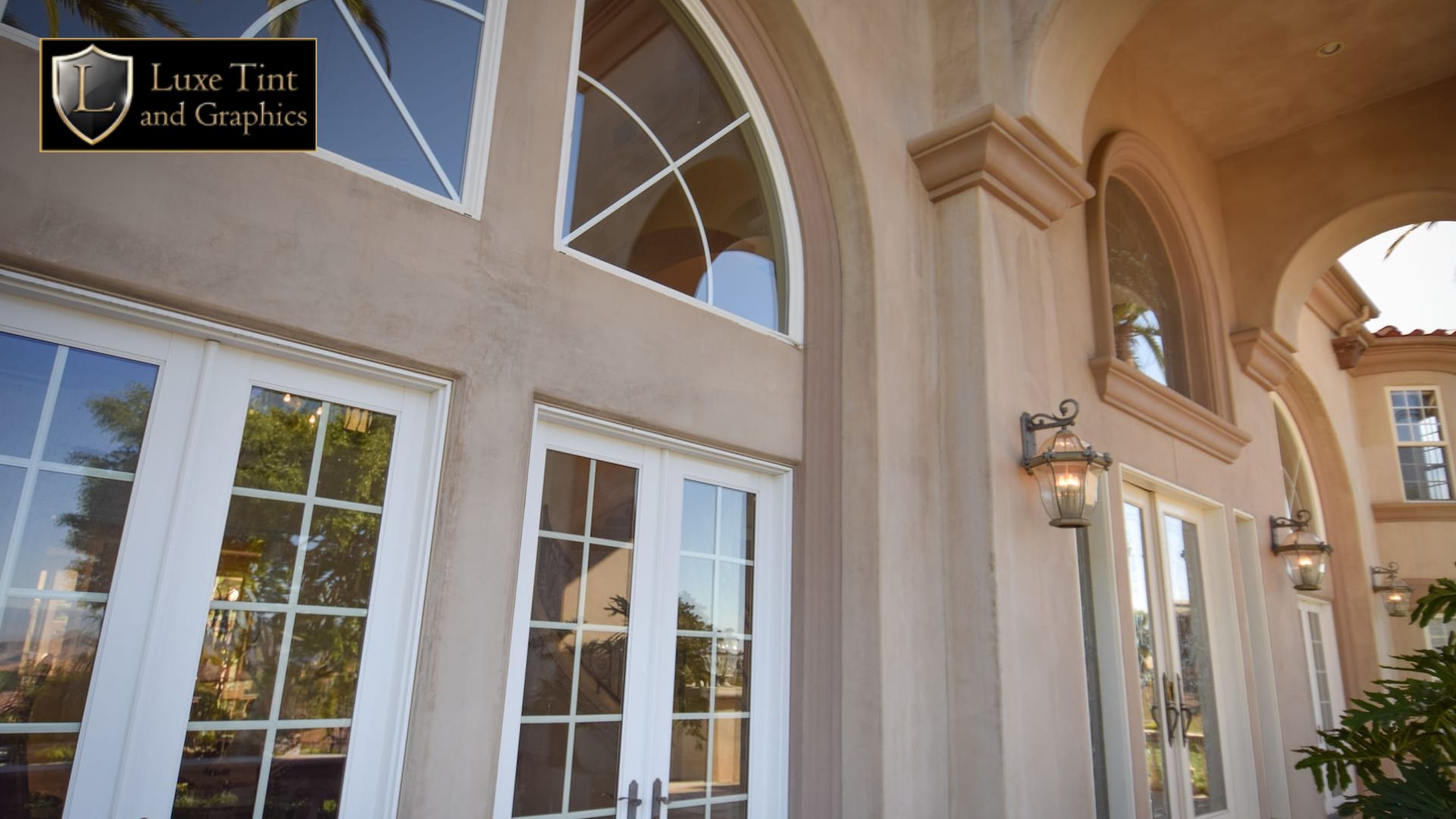 Exterior view of a luxury home with large arched windows and French doors reflecting palm trees, with the Luxe Tint and Graphics logo in the top left corner.