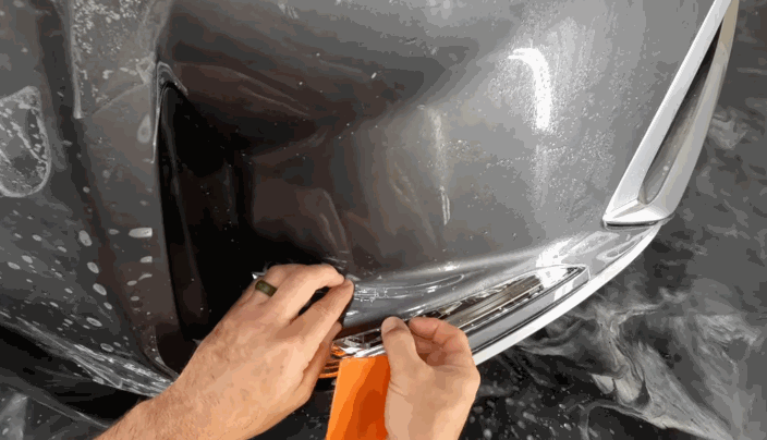 Hands stretch and tuck paint protection film around the bumper and headlight area of a gray vehicle using an orange installation tool.