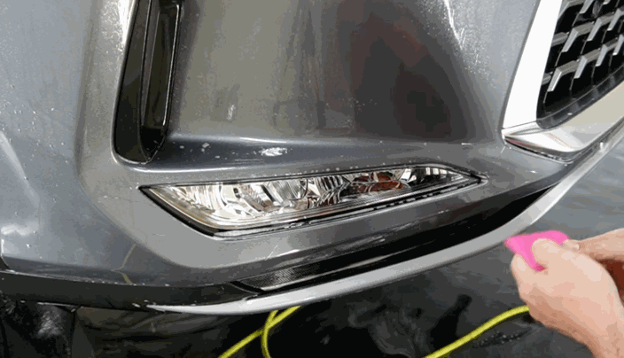 Close-up of a person applying paint protection film to a gray car bumper using a pink squeegee.