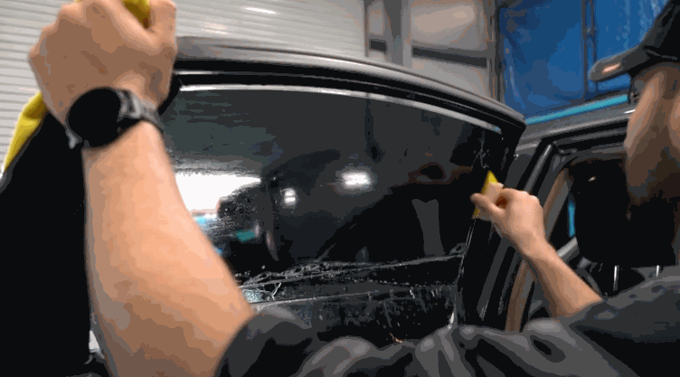 An installer smooths out window tint film on a car door window using a yellow squeegee, pressing out air bubbles and moisture.