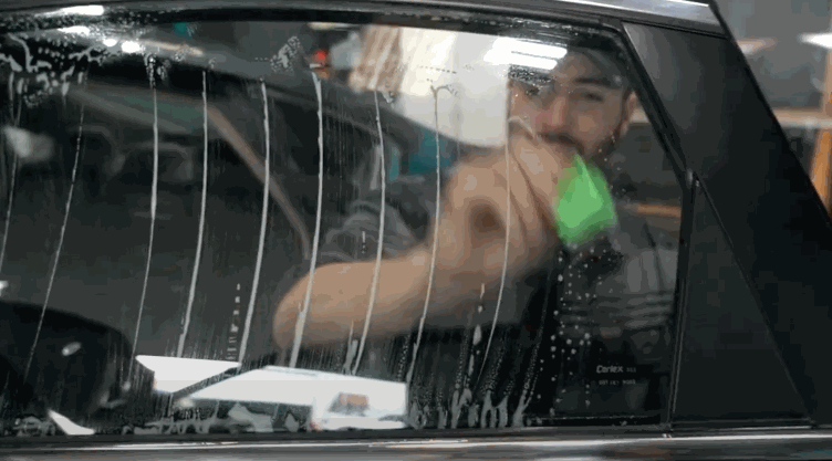 A man cleans the inside of a car window using a green scrub pad and cleaning solution, prepping the surface for window tint installation.