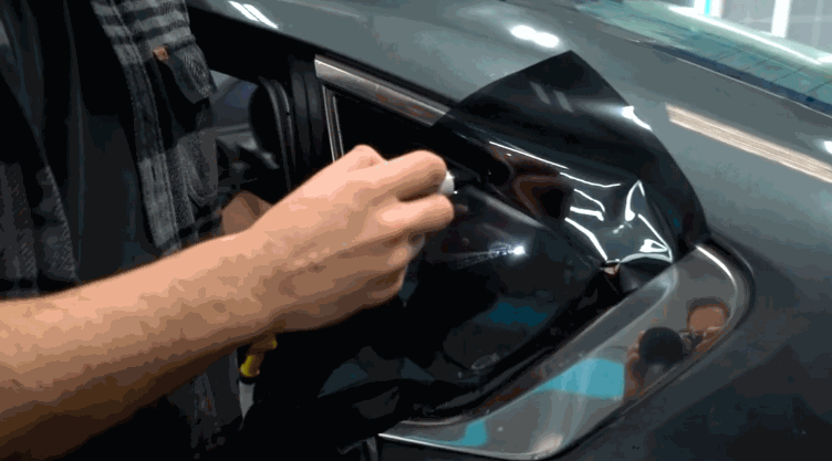 A person carefully applies dark window tint film to a small triangular rear car window using a heat gun to smooth it out.
