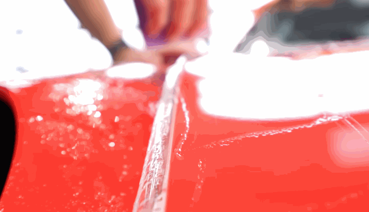 Installer uses a squeegee to apply paint protection film onto the hood of a red car with water for adhesion.
