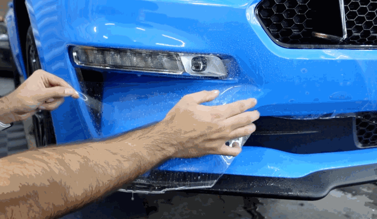 Close-up of paint protection film being carefully applied to the front bumper of a blue sports car, ensuring a seamless fit and durable finish.
