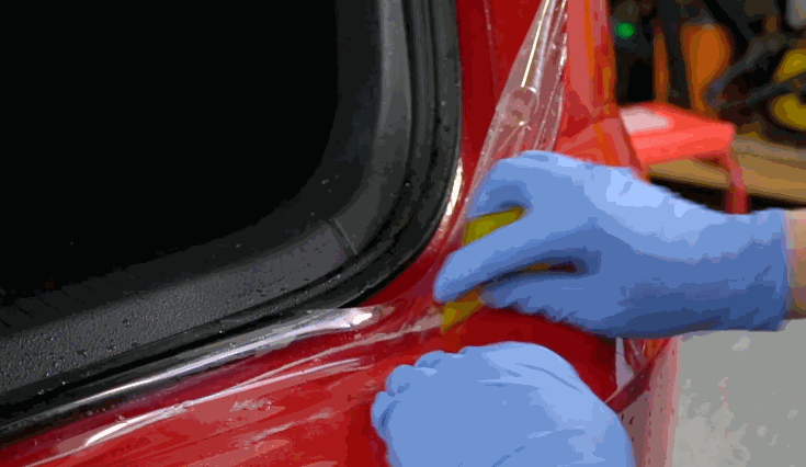 Technician in blue gloves smoothing paint protection film along the edge of a red car door using a yellow squeegee tool to ensure full coverage.