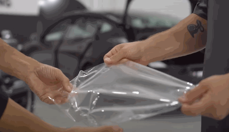 Two people stretch a section of transparent paint protection film between their hands, inspecting its flexibility before application.