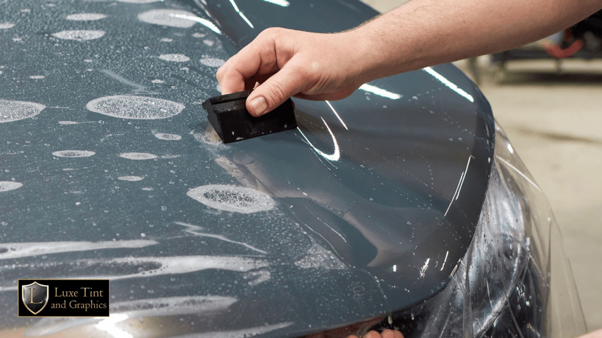 Close-up of a hand using a black squeegee to smooth out paint protection film over a soapy car hood at Luxe Tint and Graphics.